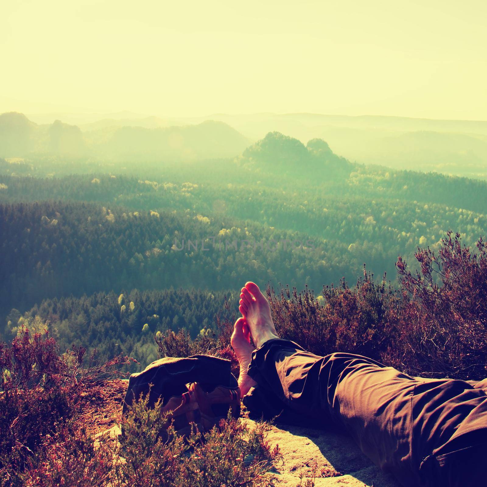 Long tired male legs in dark  hiking trousers take a rest on peak of rock above valley.  by rdonar2