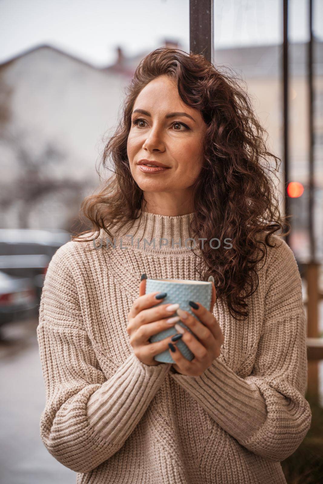 A middle-aged woman in a beige sweater with a blue mug in her hands is in a street cafe on the veranda.