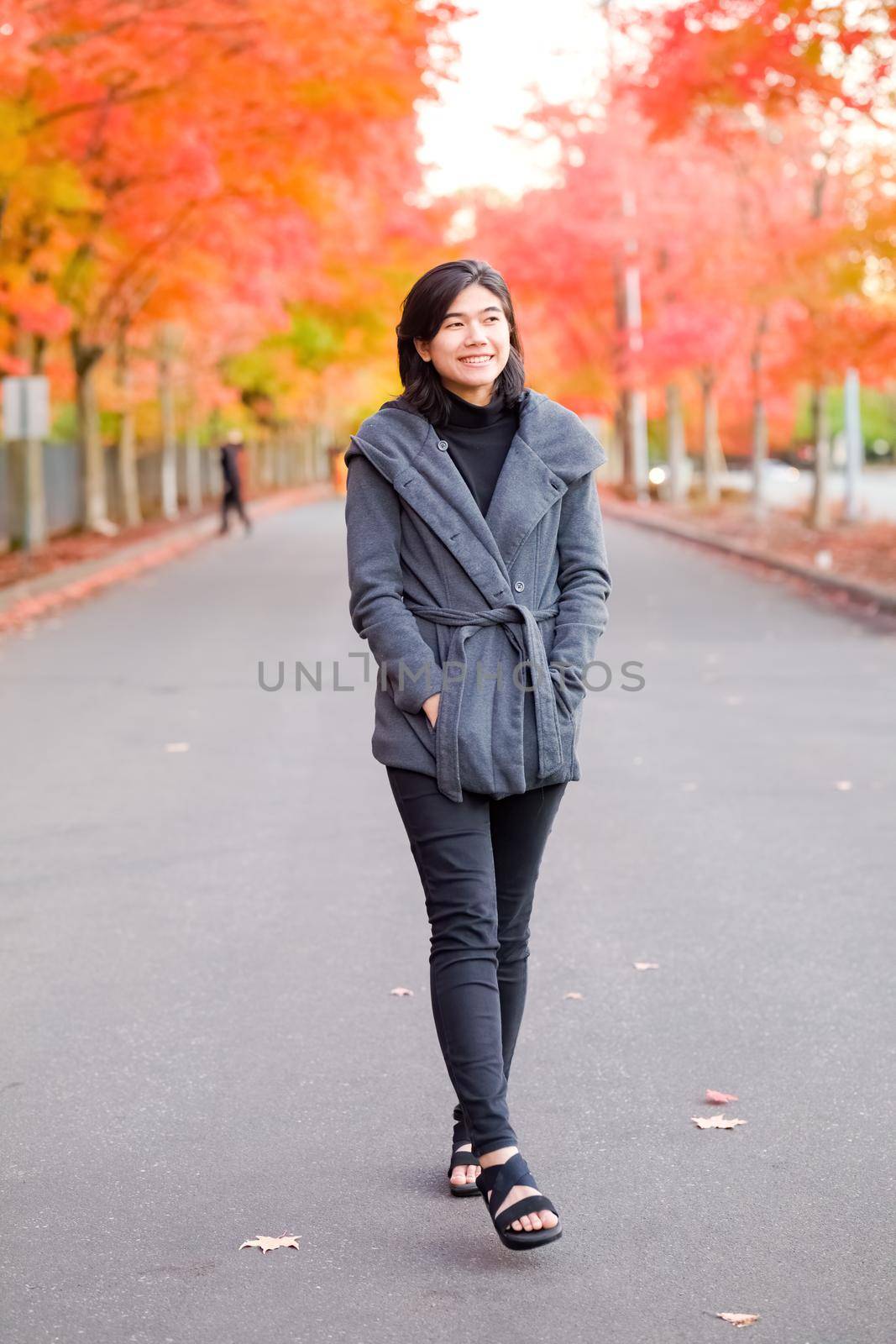 Smiling biracial teen girl or young adult female in gray jacket walking along road enjoying colorful autumn leaves