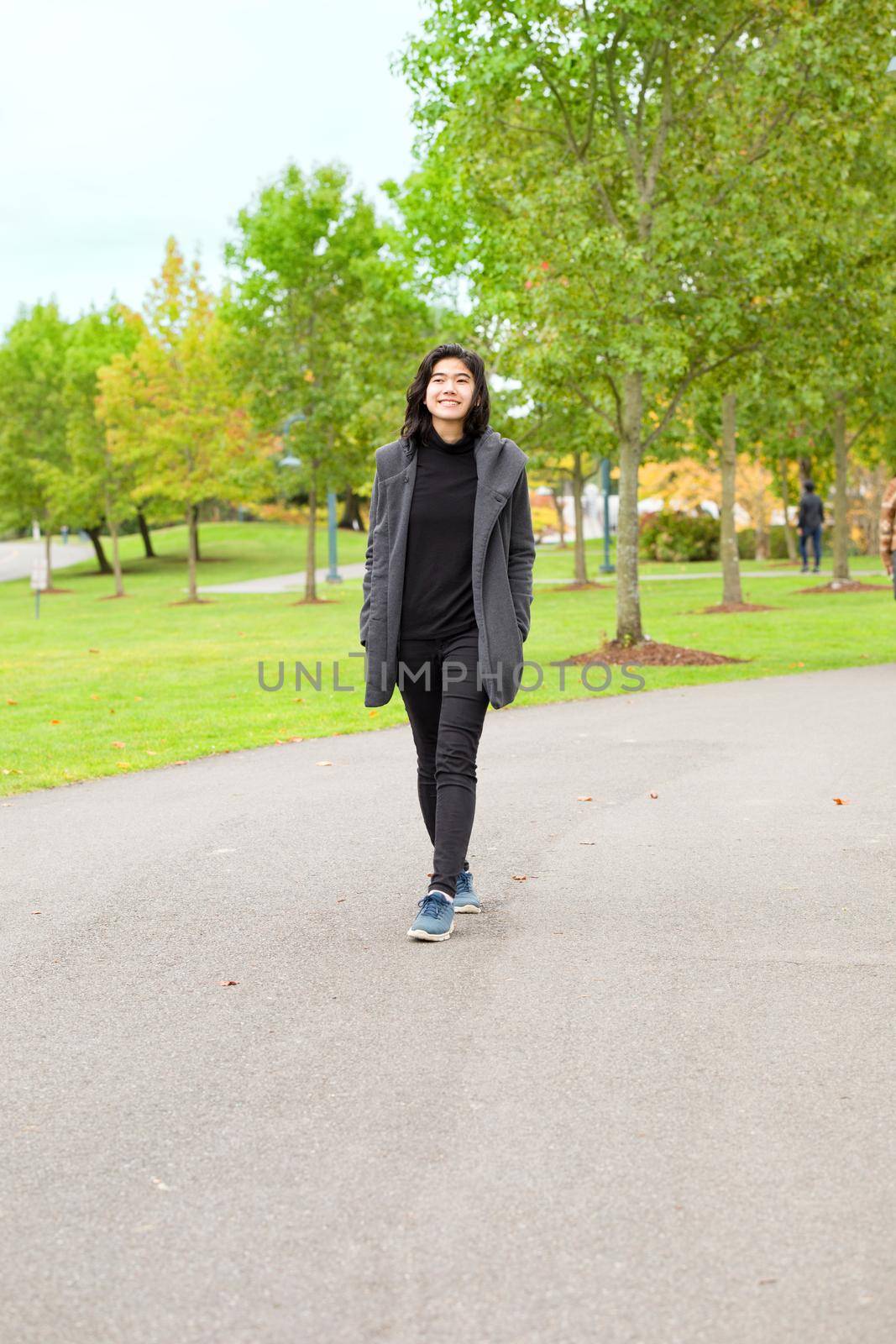 Smiling biracial teen girl or young adult female in gray jacket walking along road enjoying colorful autumn leaves