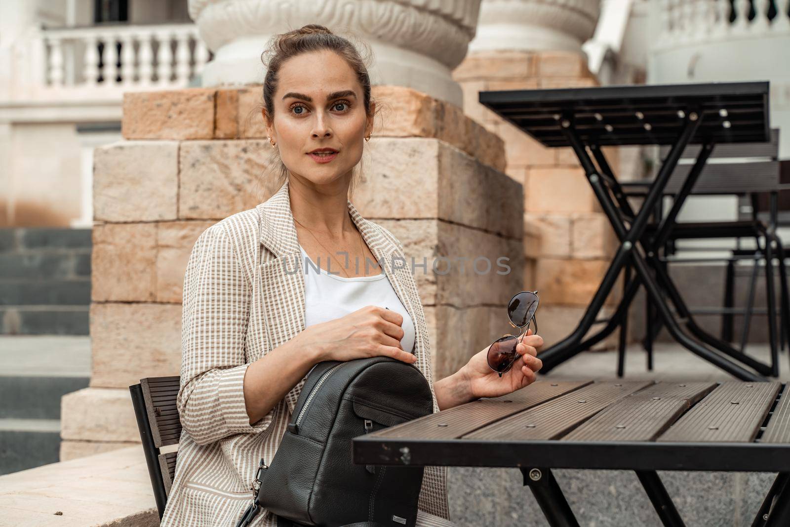 A middle aged woman sits at a table in a cafe in the hands of a black backpack. Dressed in a light suit.