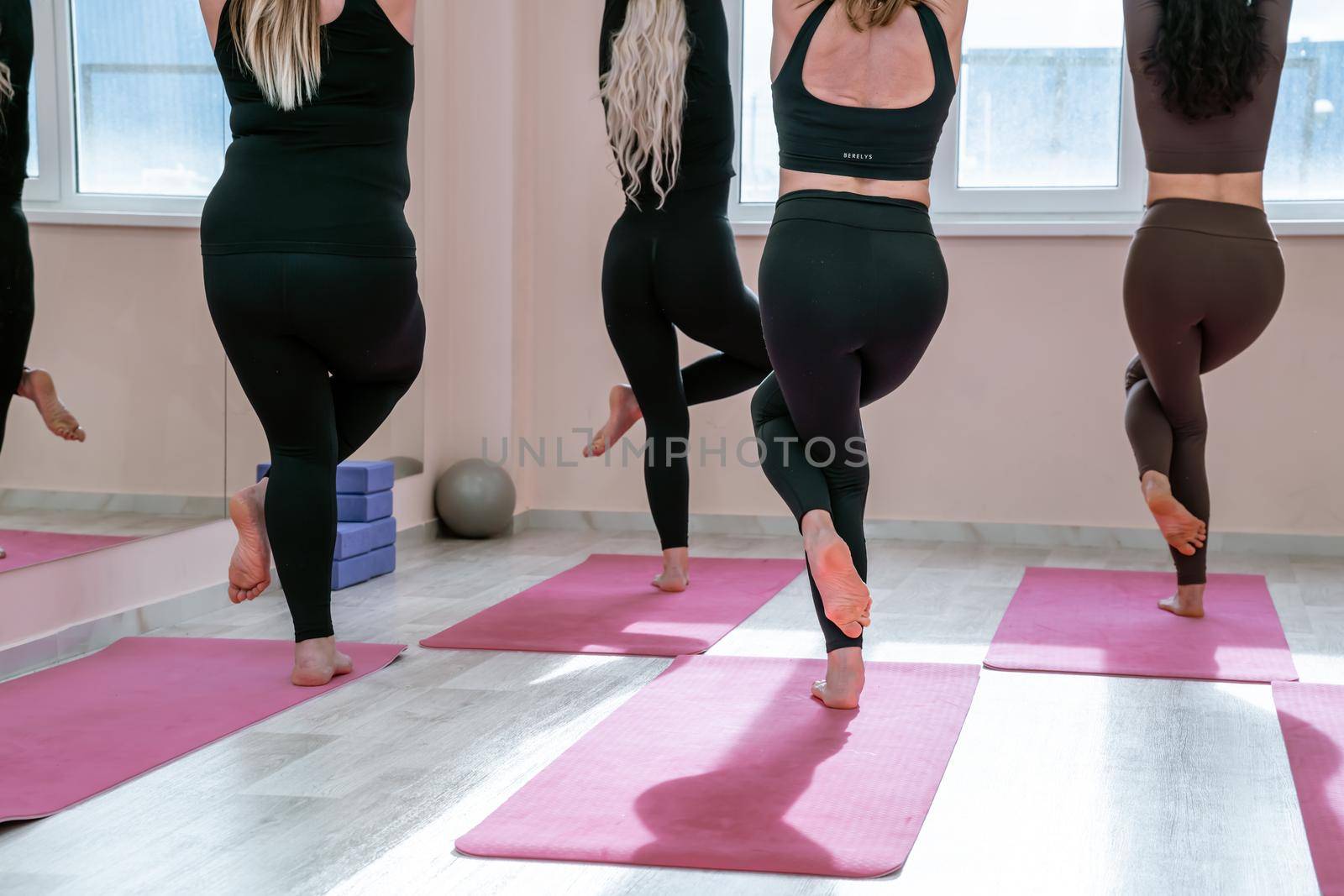 A group of six athletic women doing pilates or yoga on pink mats in front of a window in a beige loft studio interior. Teamwork, good mood and healthy lifestyle concept