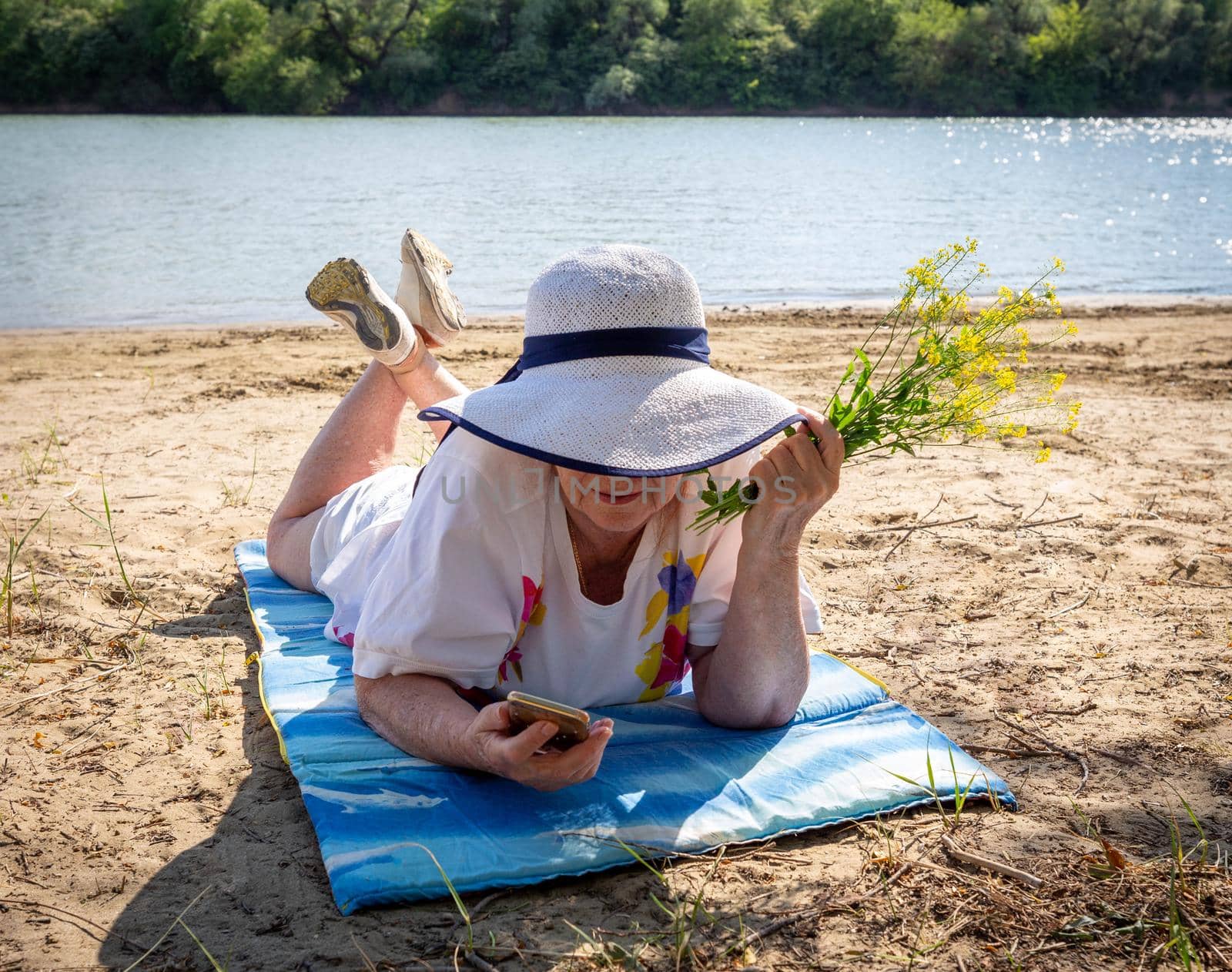 Pensive elderly woman, elderly pensioner, rest on the sand by the river, the concept of time and age, summer vacation in the fresh air, healthy lifestyle