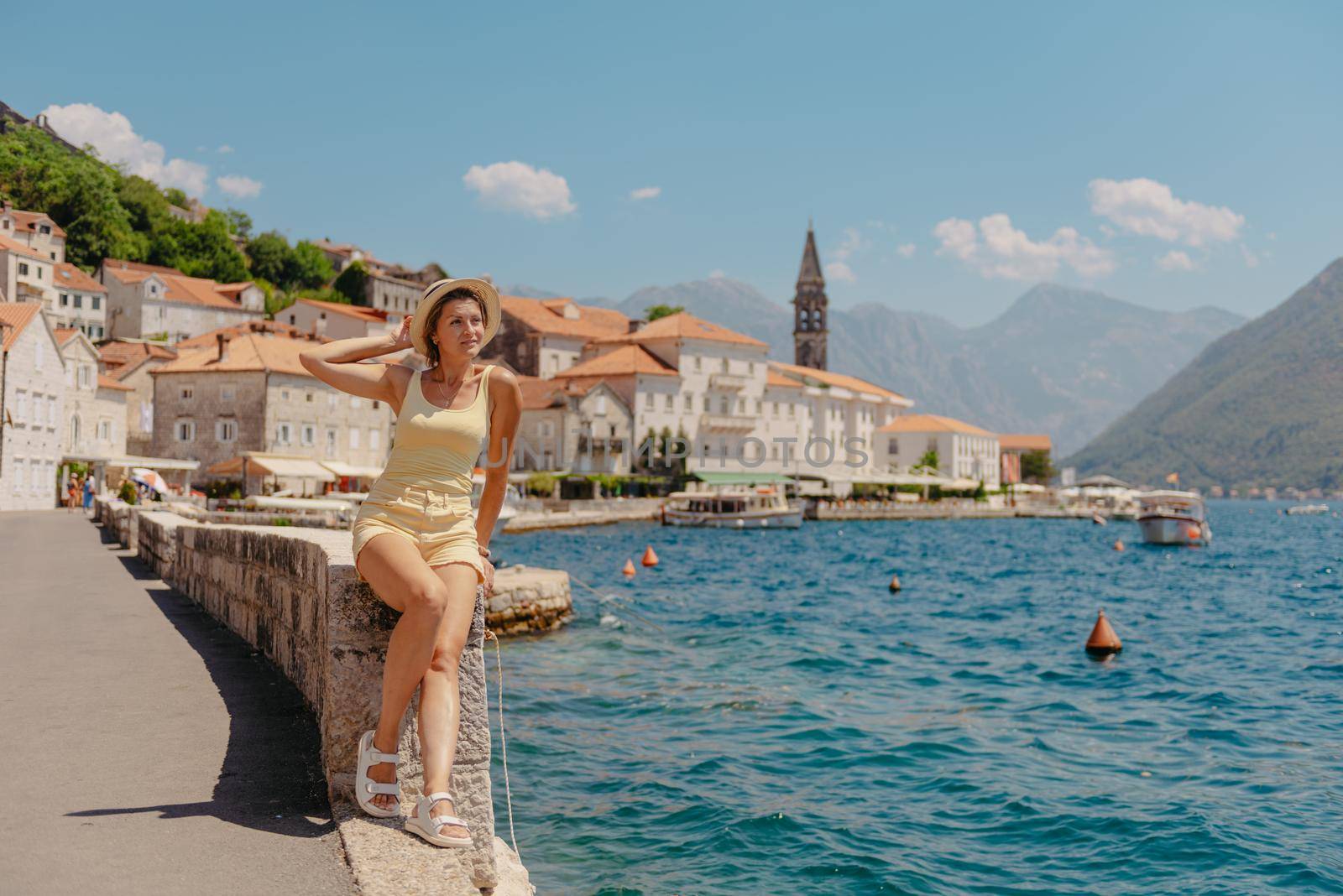 Summer photo shoot on the streets of Kotor, Montenegro. Beautiful girl in yellow dress and hat. smiling tourist girl with hat. Spectacular view of Montenegro with copy space. Ю fashion outdoor photo of beautiful sensual woman with blond hair in elegant dress and straw hat and bag, posing in Montenego's city Perast.