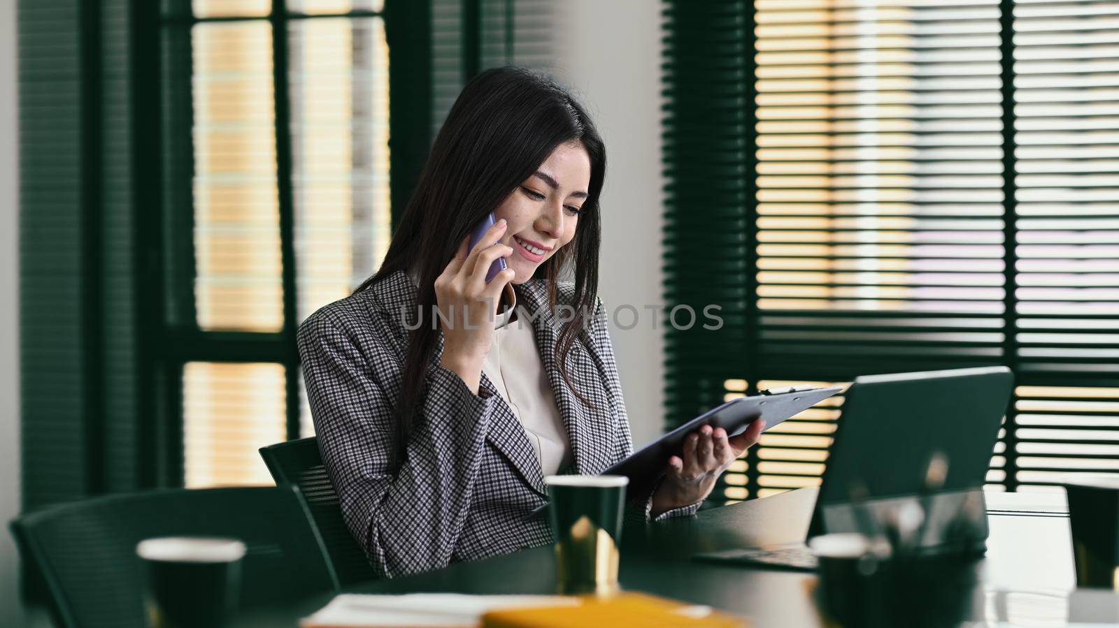 Millennial businesswoman in formal suit looking at document and and talking on mobile phone with business partner.
