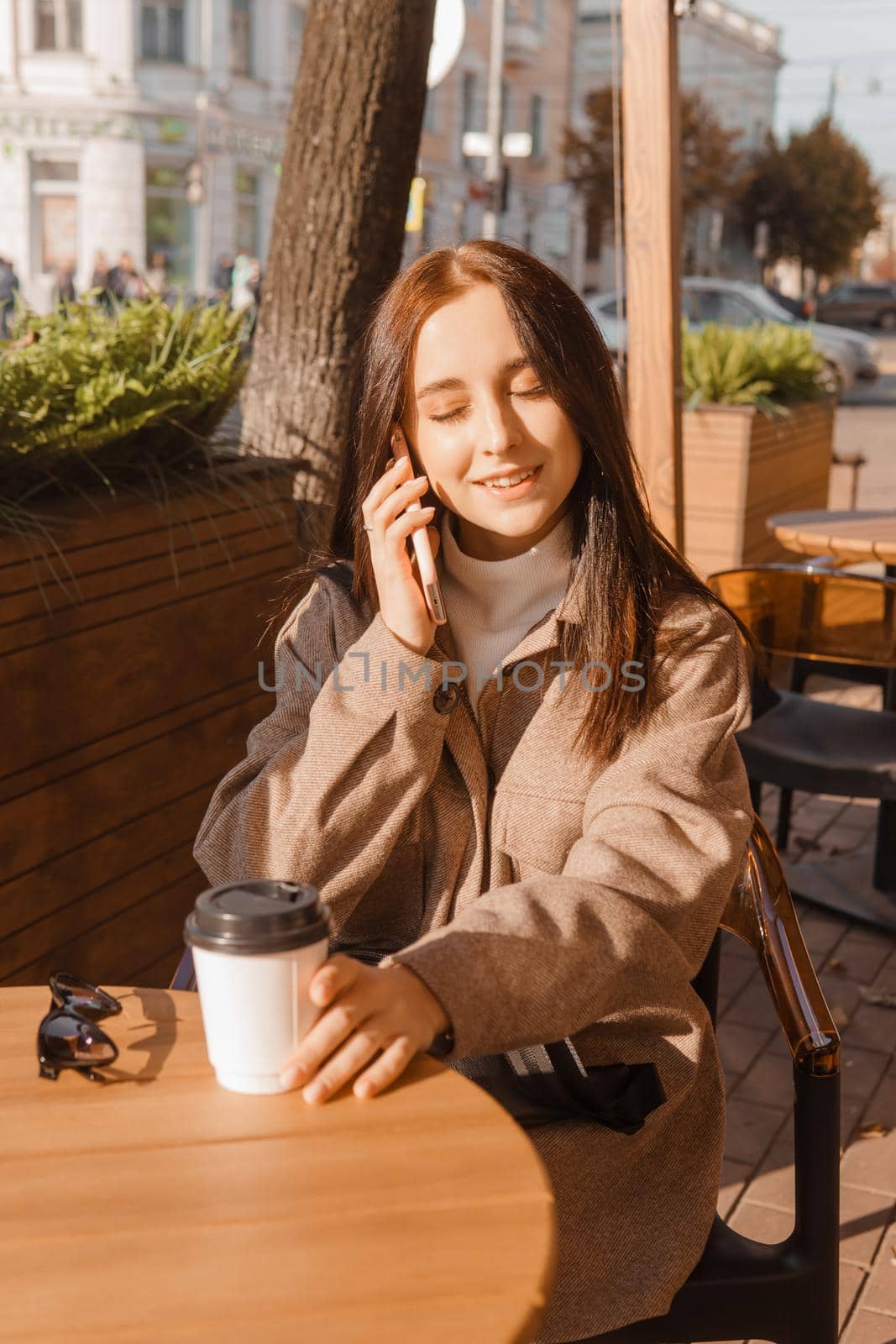 A stylish brunette woman is sitting at a table in an outdoor cafe with coffee and a phone. The girl is talking on the phone. Autumn walk.