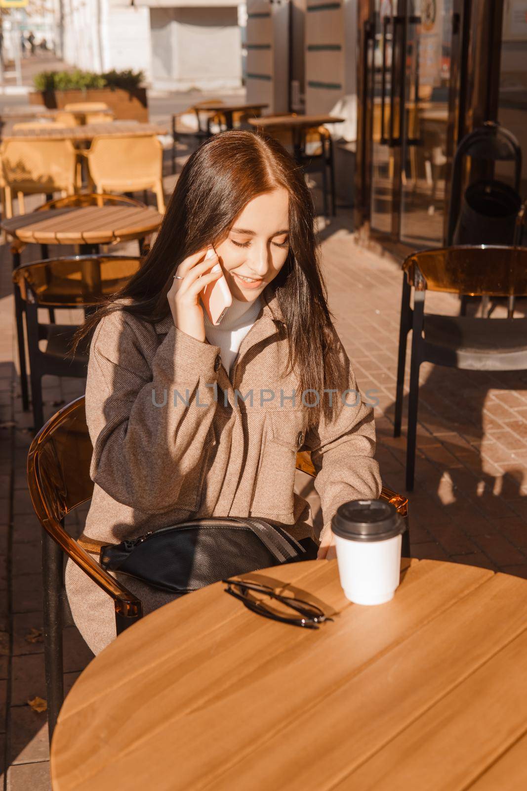 A stylish brunette woman is sitting at a table in an outdoor cafe with coffee and a phone. The girl is talking on the phone. Autumn walk.