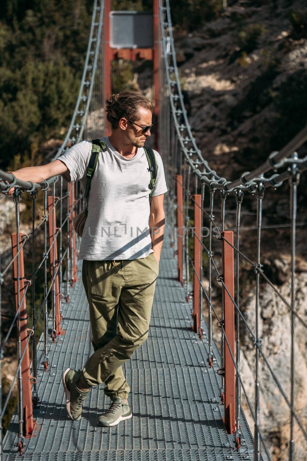 A man with a backpack on a suspension bridge against the backdrop of a mountain landscape with trees. Hiking trail pass through the hanging bridge. Travel and exploration concept.