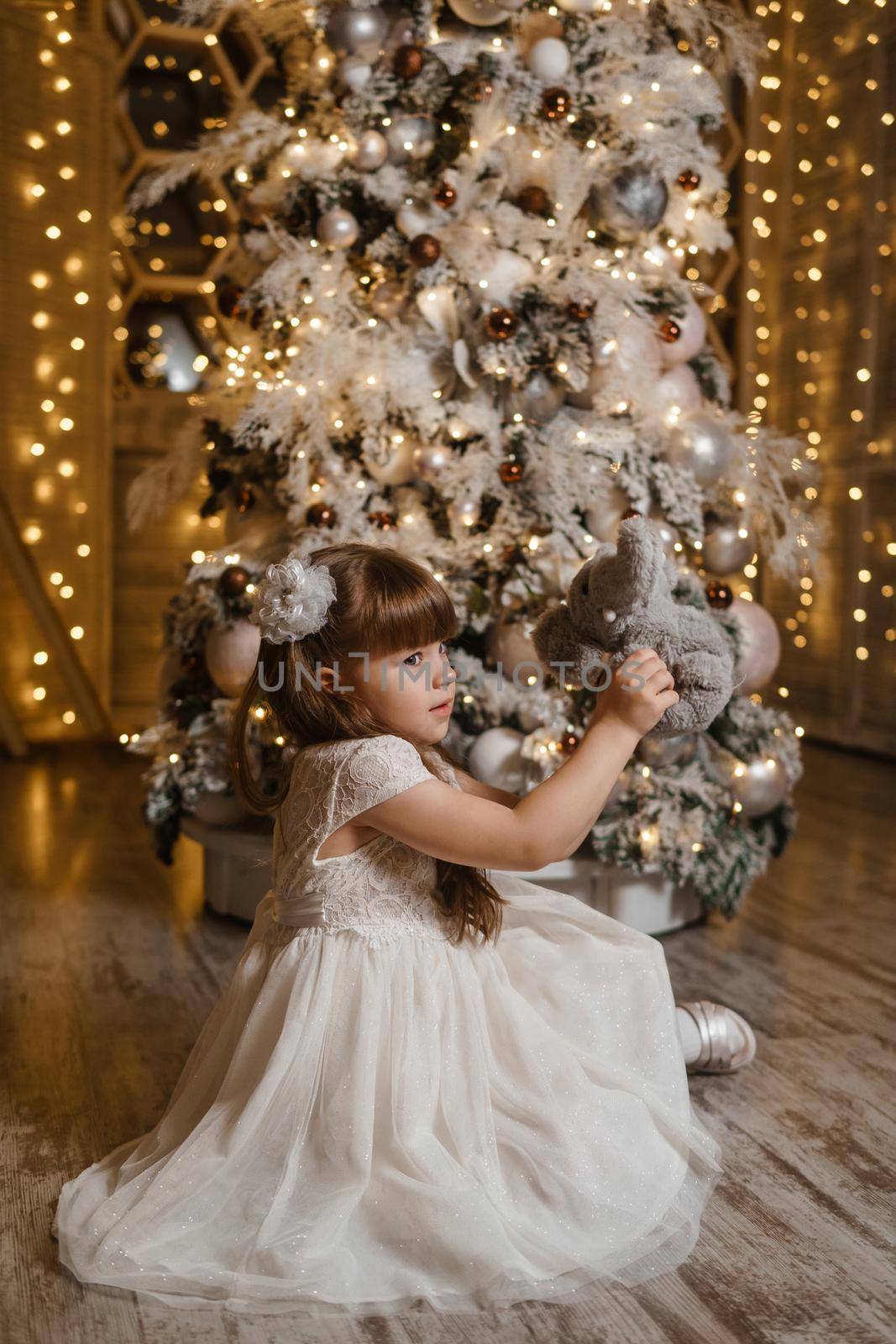 A girl in a festive light dress next to a Christmas tree, lights of garlands in the background. The concept of New Year holidays.
