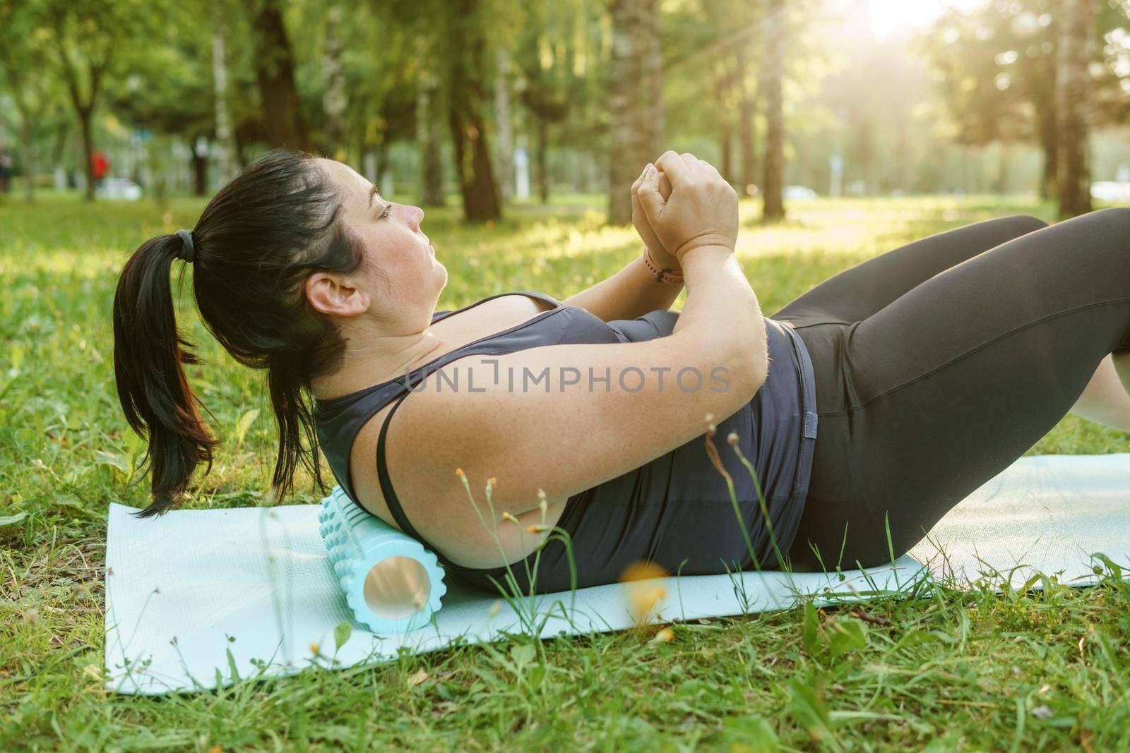 A charming brunette woman plus-size body positive practices sports in nature. Woman is engaged with a massage roller for the body in the park on a sports mat.