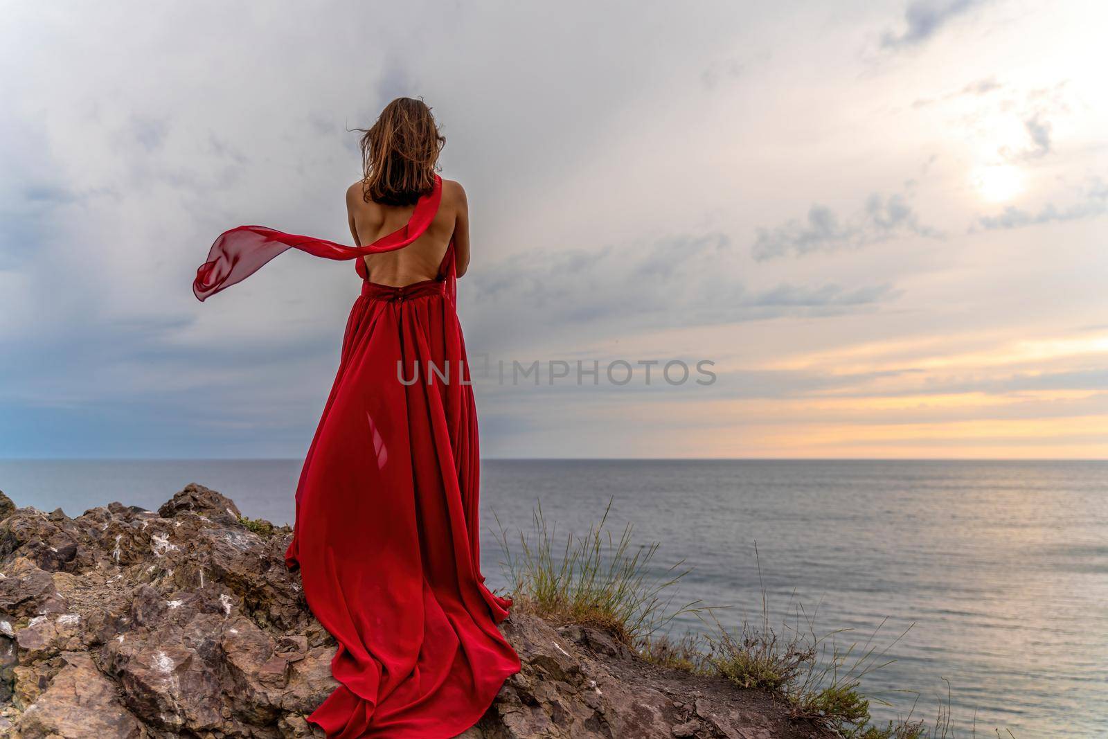 A woman in a red dress stands above a stormy sky, her dress fluttering, the fabric flying in the wind