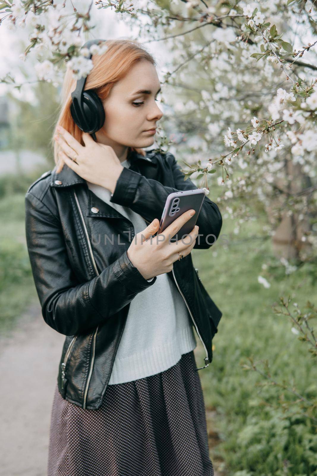 A red-haired teenage girl listens to music in headphones, configured via the phone. The time of spring flowering of cherry orchards.