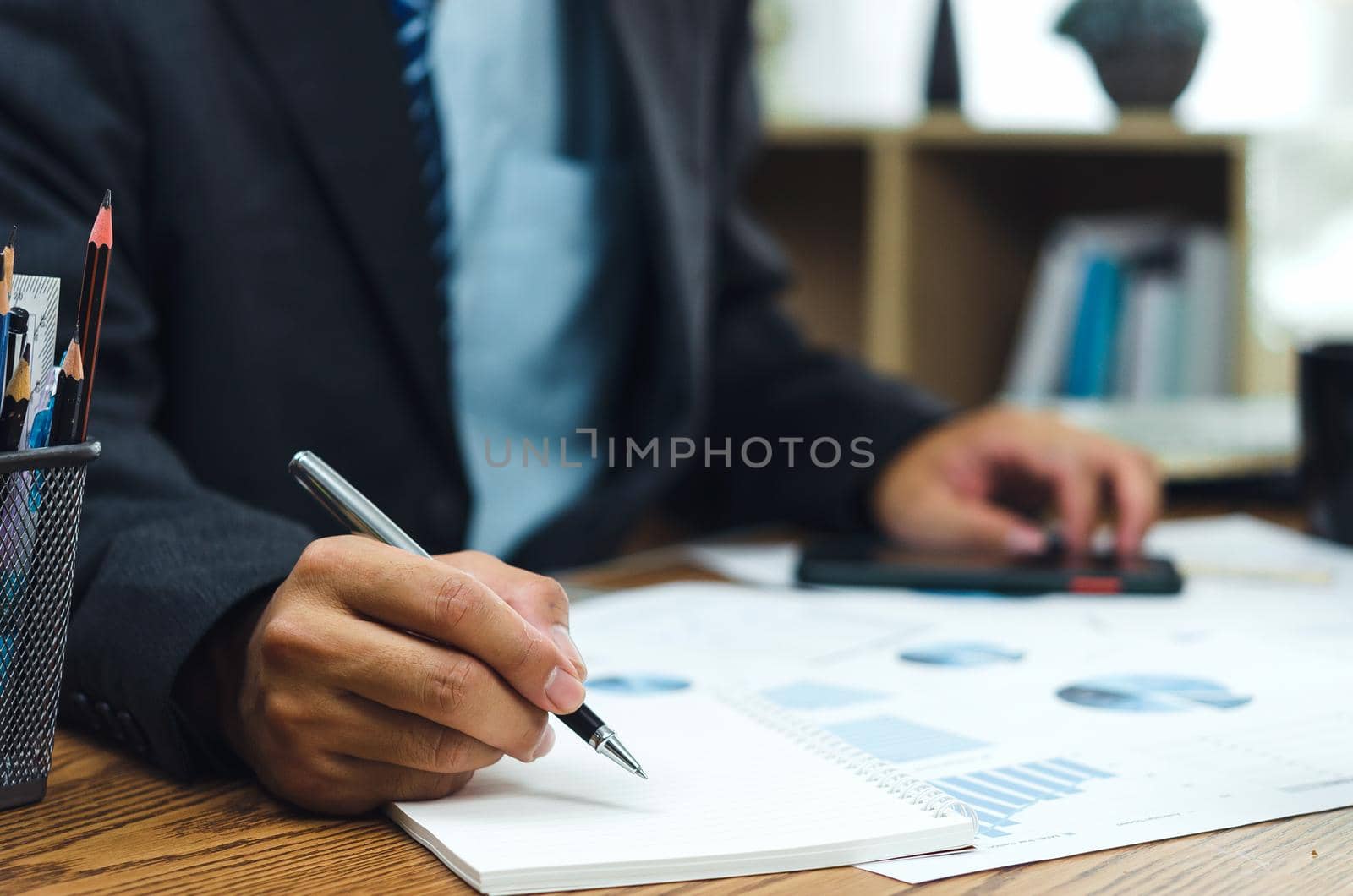 Businessman holding pen writing a book and document graph accounting finance paper work analysis chart marketing investment and report economy on desk at office.