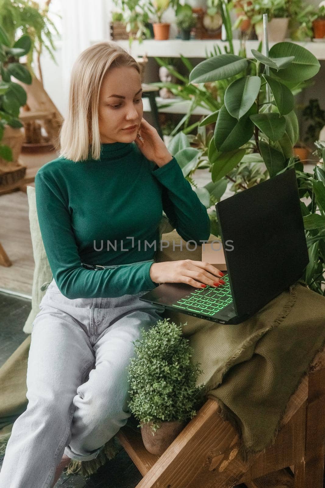 A blonde woman in a room with a lot of green indoor plants is working on a laptop. The concept of biophysical design in the interior. Work from home, work as a freelancer.