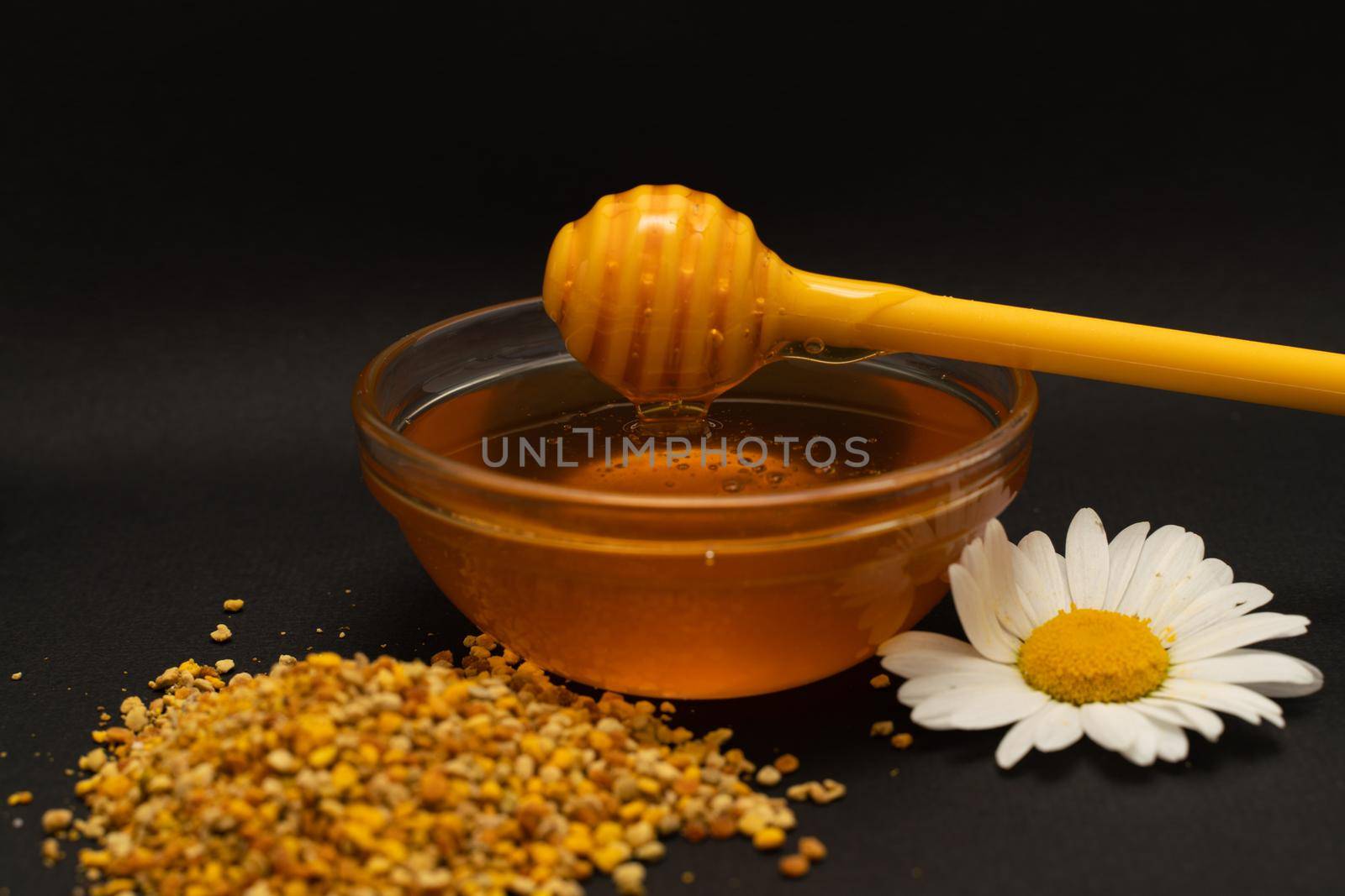 A yellow spoon of honey dipped in a jar of honey and bee bread scattered on a black background decorated with live chamomile. Honey dripping around, nice and inviting photos. Healthy food concept
