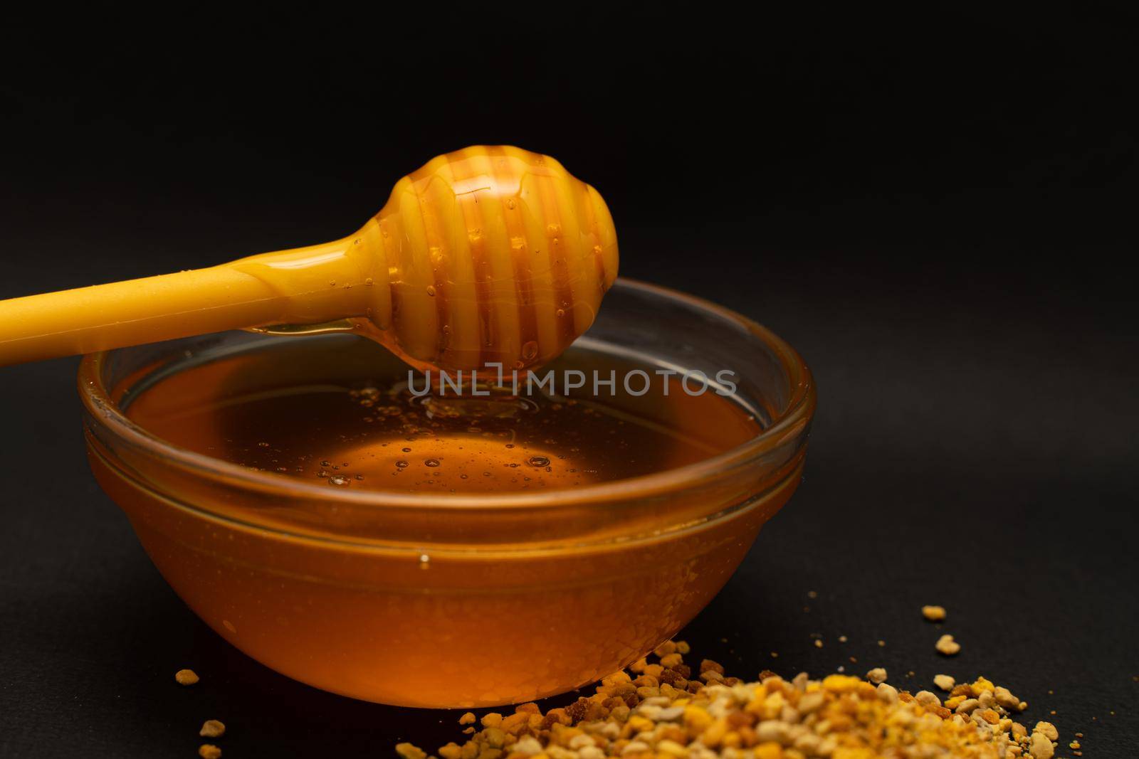A yellow spoon of honey dipped in a jar of honey and bee bread scattered on a black background. Honey dripping around, nice and inviting photos. Healthy food concept