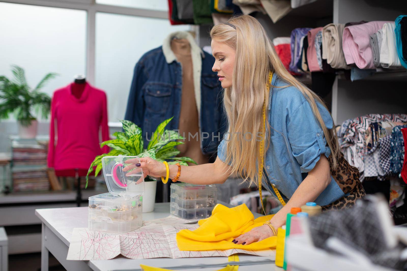 A woman reaches into containers of small ornaments that are on a tailor's table. Working in a tailor's workshop. A blonde woman with long hair.