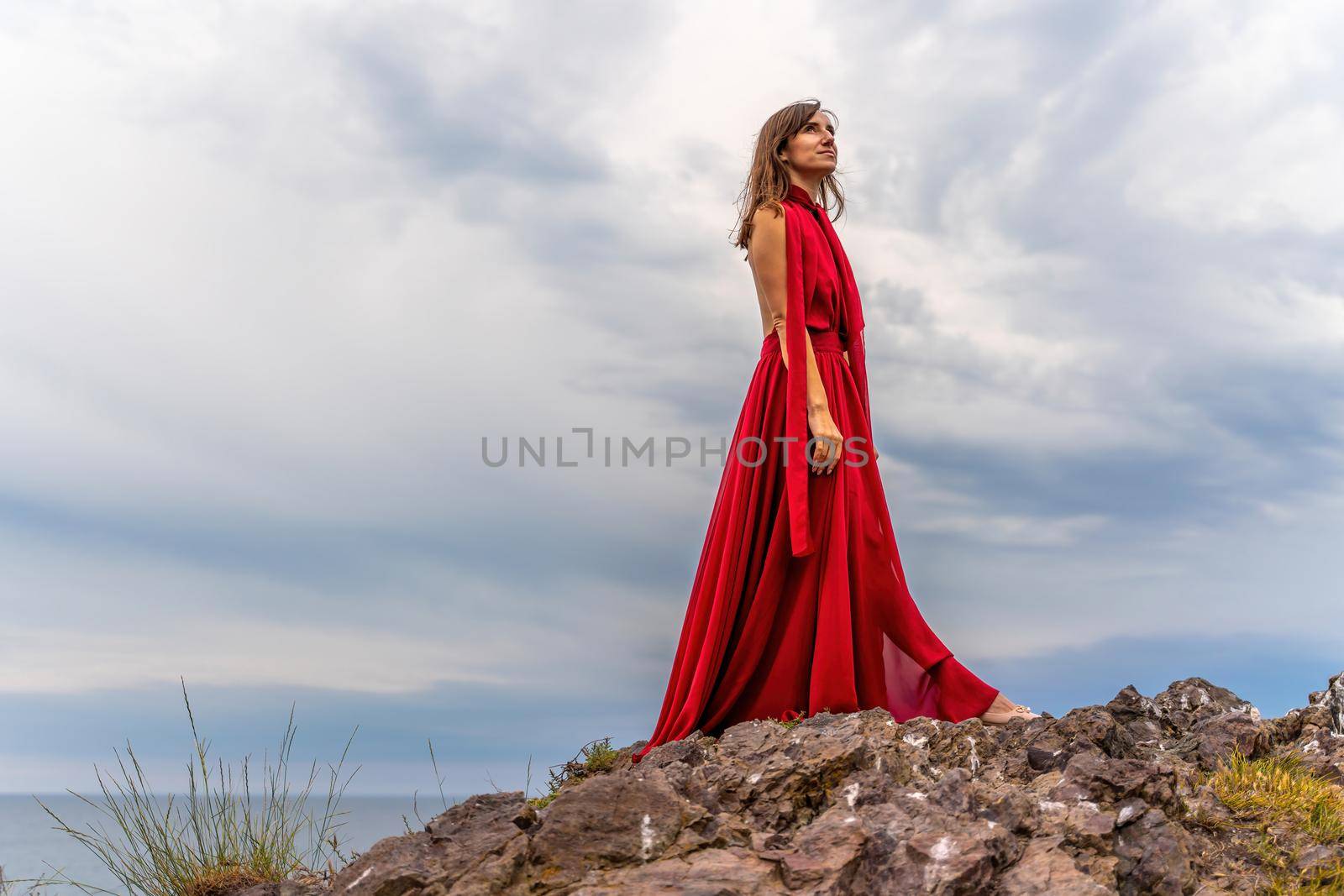 A woman in a red dress stands above a stormy sky, her dress fluttering, the fabric flying in the wind