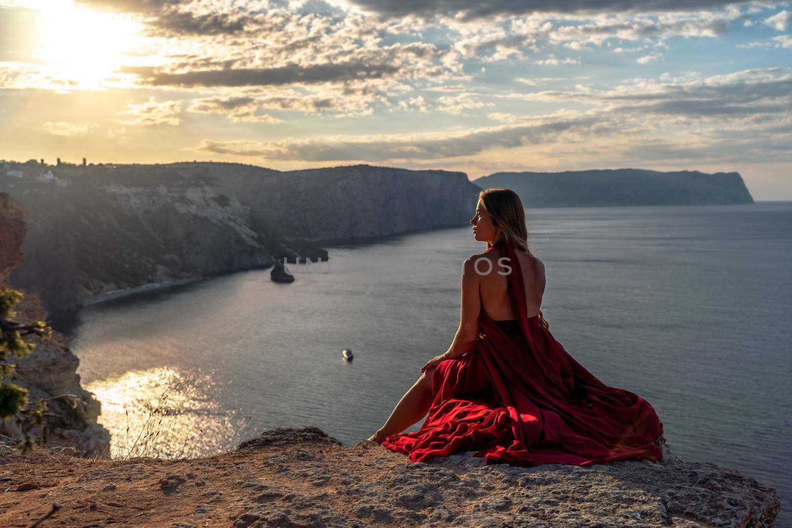 A girl with loose hair in a red dress sits on a rock rock above the sea. In the background, the sun rises from behind the mountains. by Matiunina
