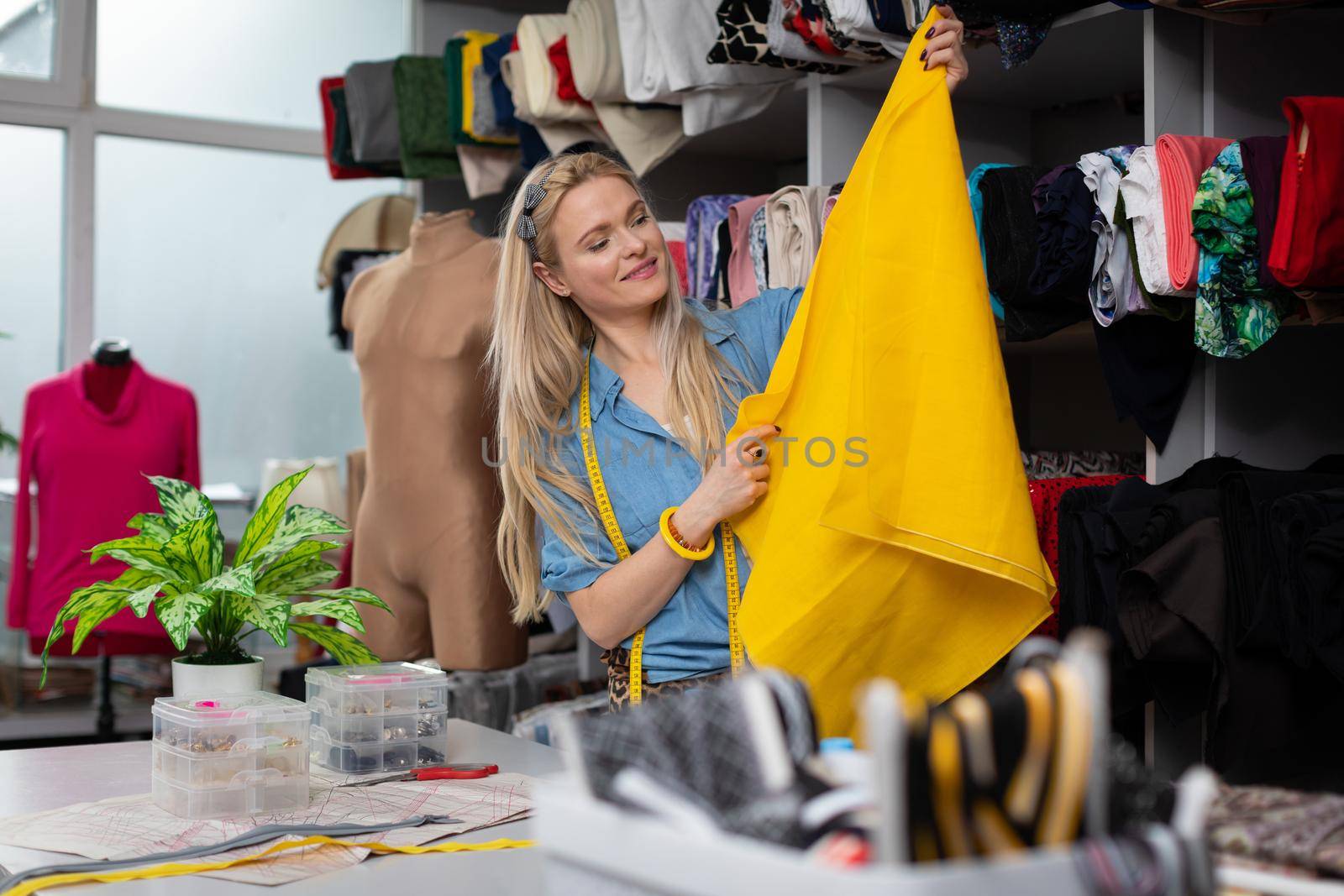 A blonde woman lifts up a piece of yellow fabric and checks in the light how the color looks. Working in a tailor's shop. She inspects the fabric.