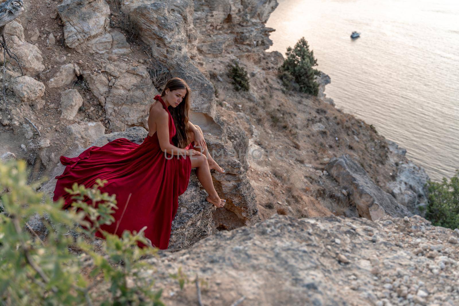 A girl with loose hair in a red dress sits on a rock rock above the sea. In the background, the sea. The concept of travel. by Matiunina