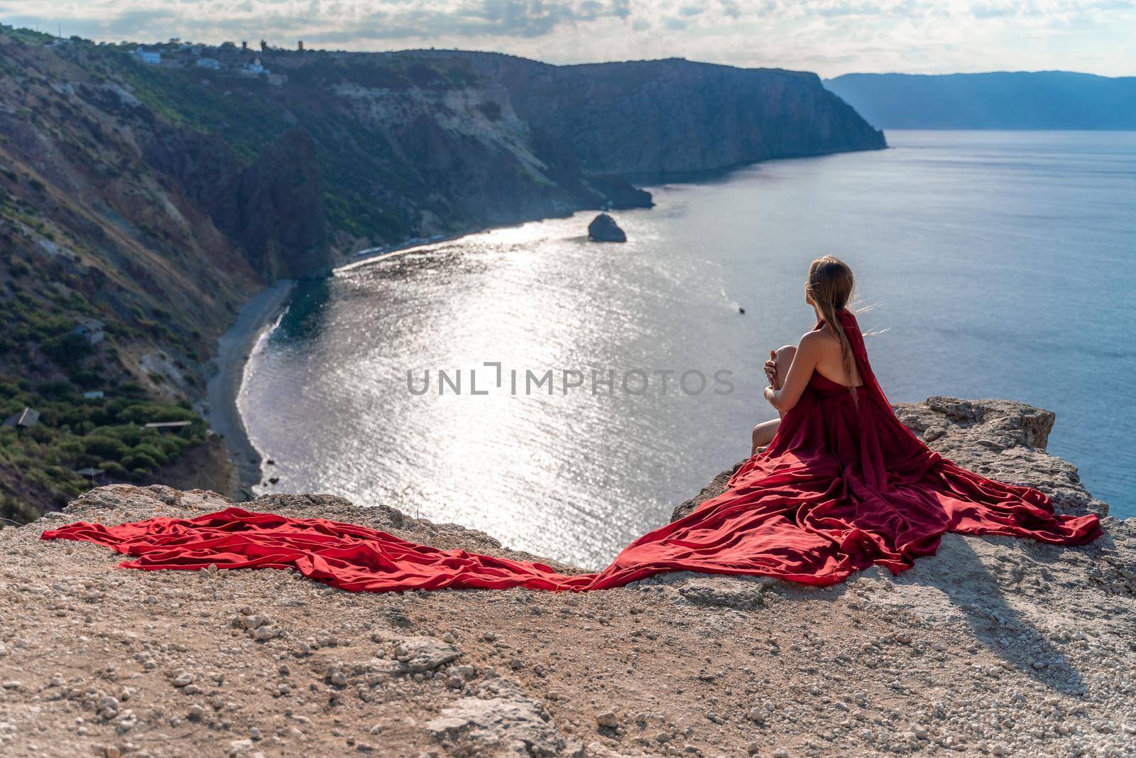 A girl with loose hair in a red dress sits on a rock rock above the sea. In the background, the sun rises from behind the mountains. by Matiunina