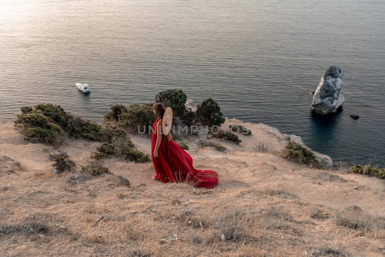 A girl with loose hair in a red dress and hat stands on the rocks above the sea. In the background, the sea and the rocks. The concept of travel. by Matiunina