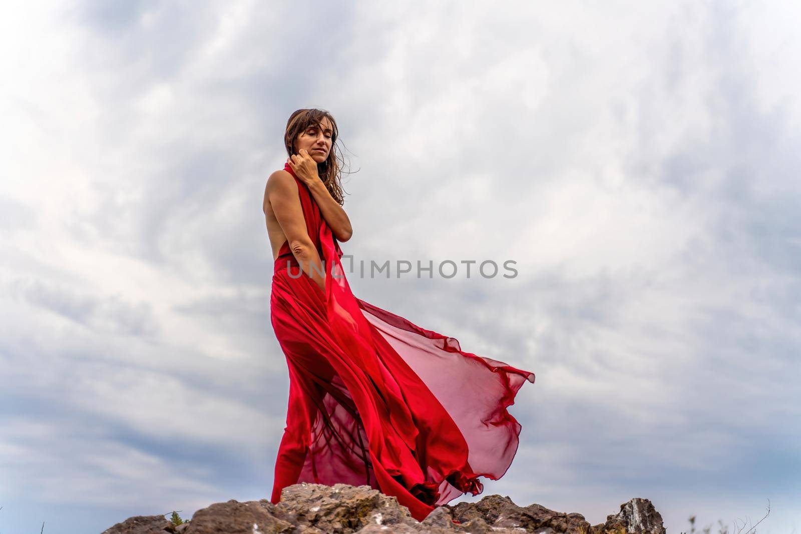 A woman in a red dress stands above a stormy sky, her dress fluttering, the fabric flying in the wind