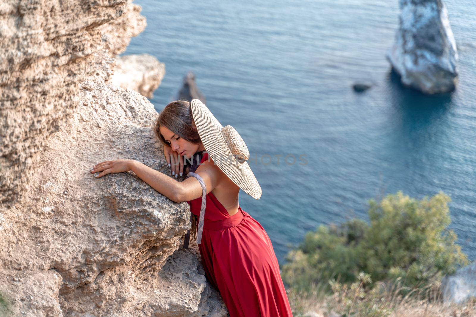 A girl with loose hair in a red dress and hat stands on the rocks above the sea. In the background, the sea and the rocks. The concept of travel