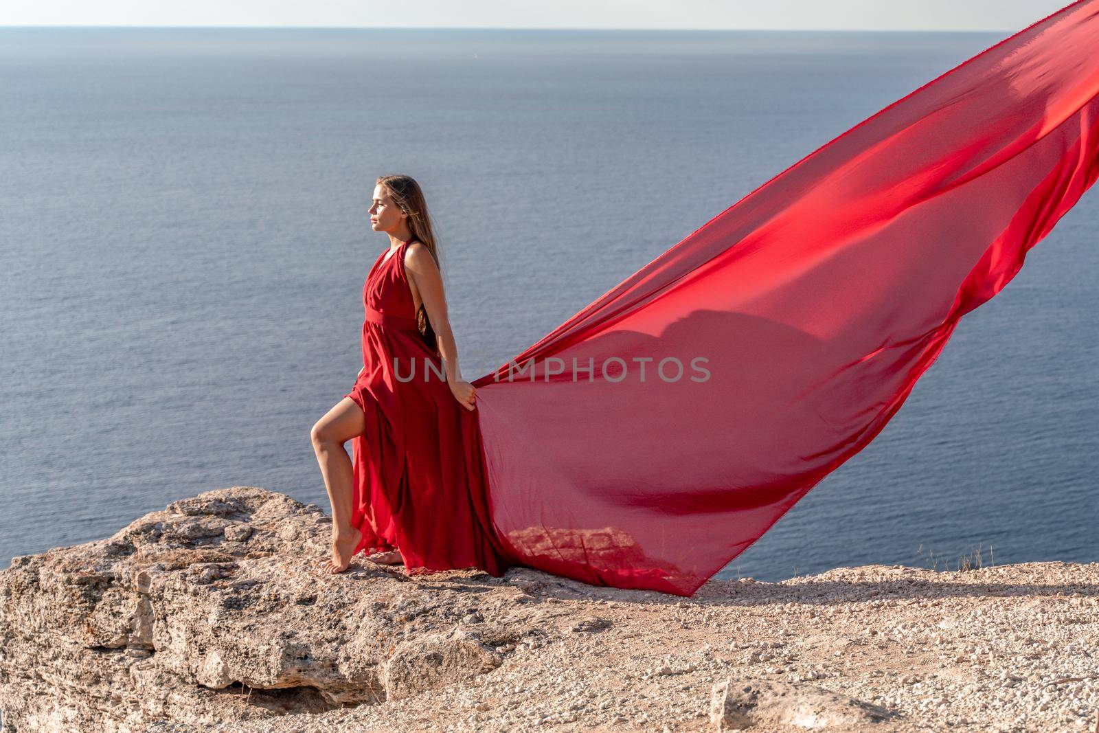 A girl with loose hair in a red dress stands on a rock rock above the sea. In the background, the sea and the rocks. The concept of travel.