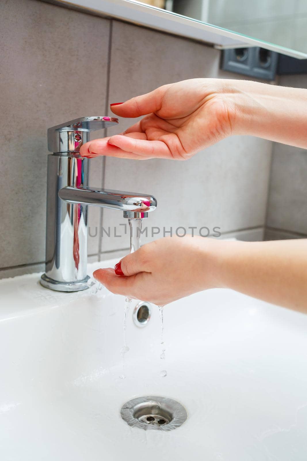 White sink with a silver faucet in the bathroom. Gray can with liquid soap for hands. Turning on the tap water, personal hand hygiene. Hand washing under running water