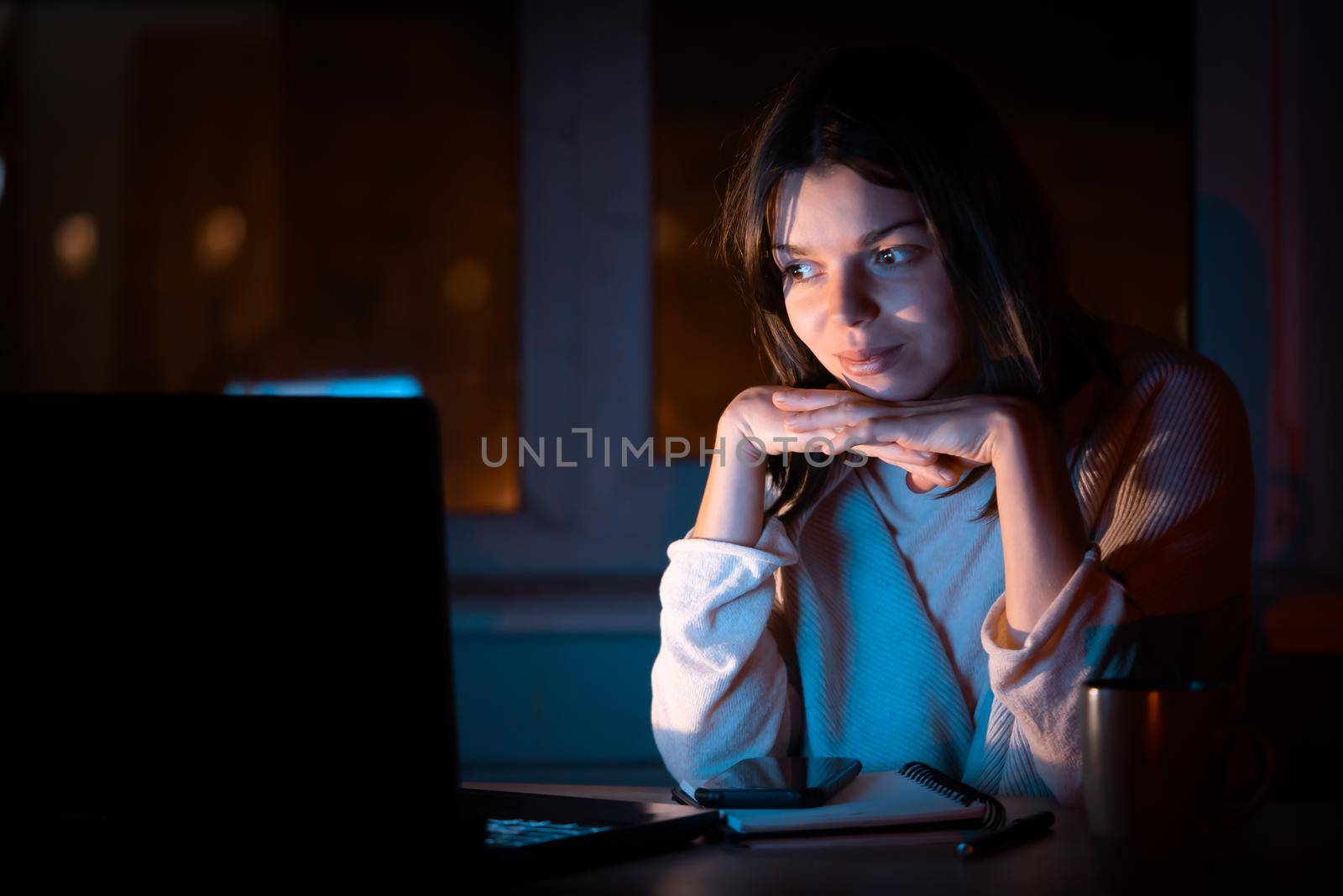 A young girl works at home at night with a laptop online, a woman with a smiley face is studying, looking for information, using her device