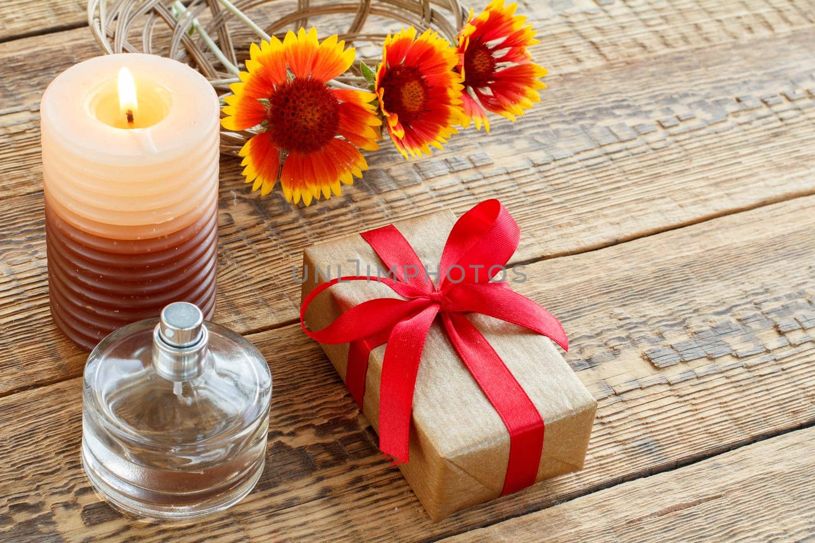 Perfume, gift or present box wrapped in kraft paper with red ribbon, burning candle and yellow flowers on wooden boards. Top view. Holiday concept.