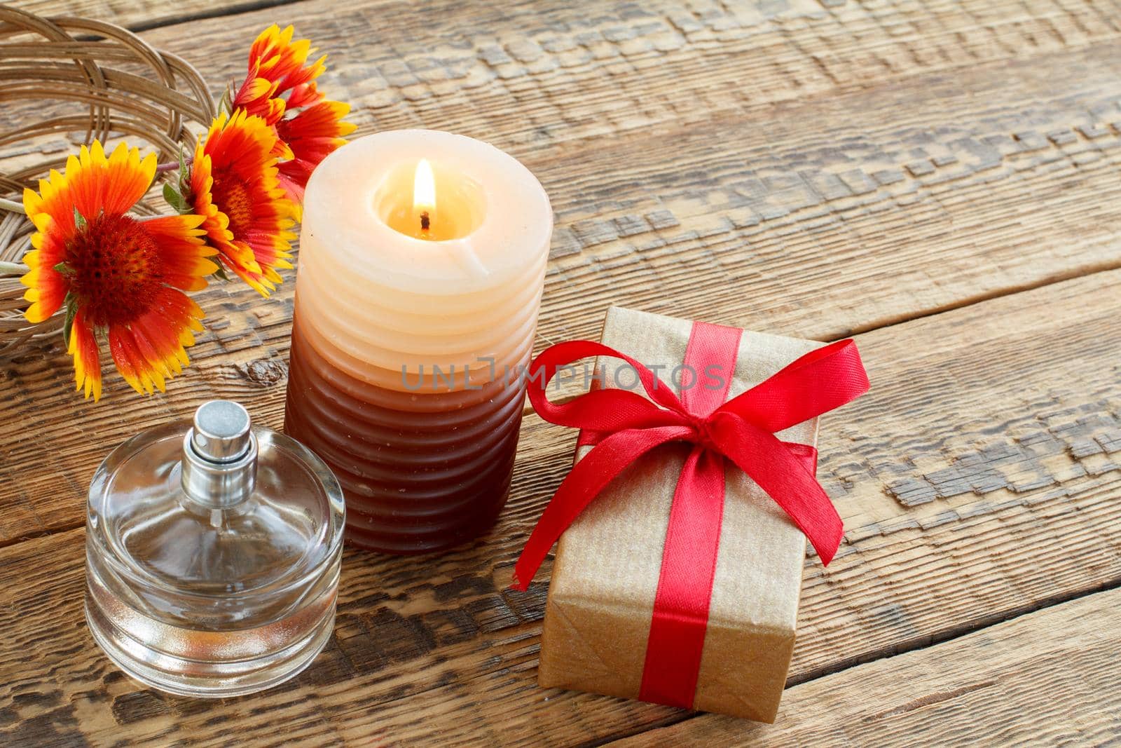 Perfume, gift box wrapped in kraft paper with red ribbon, burning candle and flowers on wooden boards. Top view. Holiday concept.