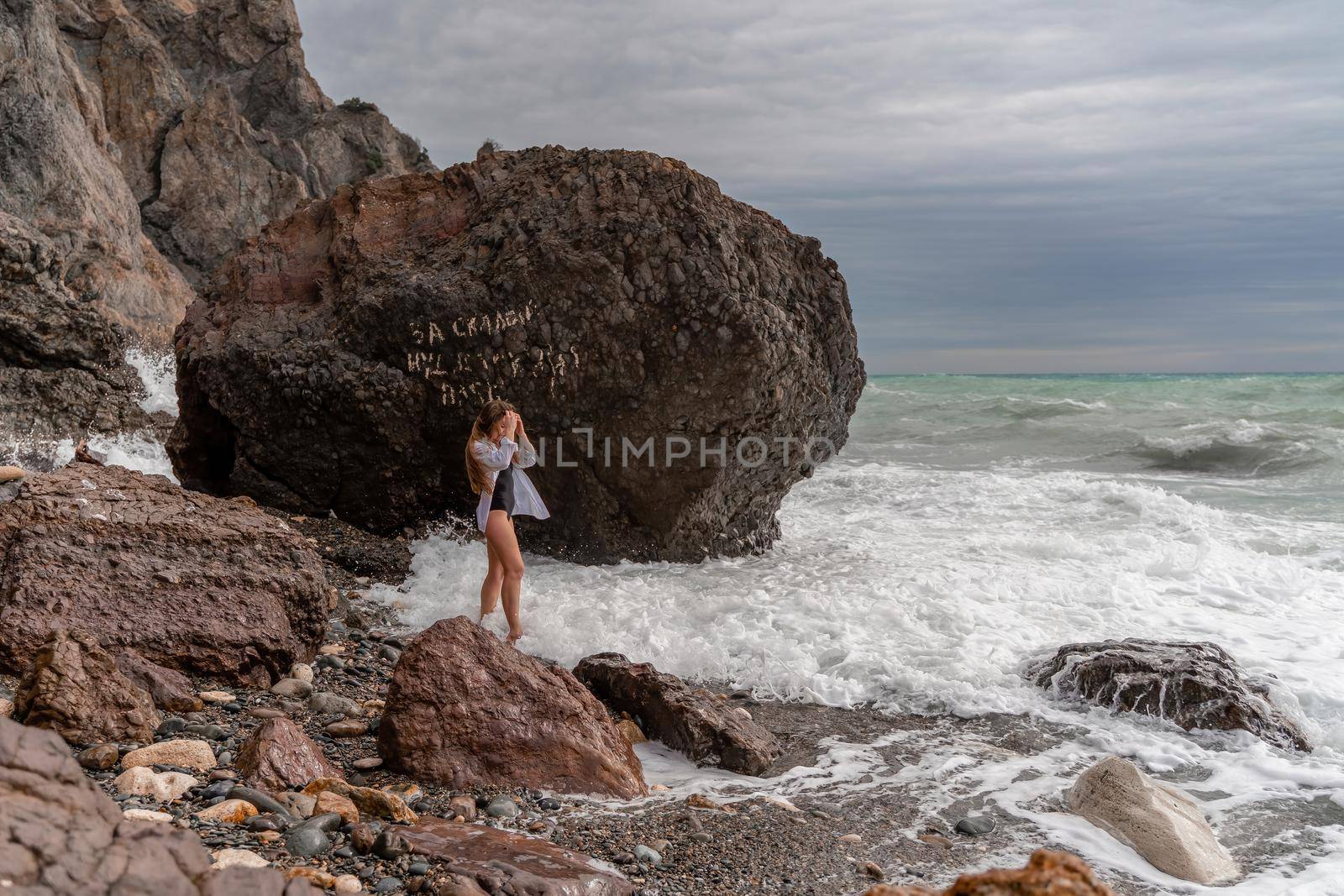 A beautiful girl in a black dress is walking on the waves, big waves with white foam. A cloudy stormy day at sea, with clouds and big waves hitting the rocks