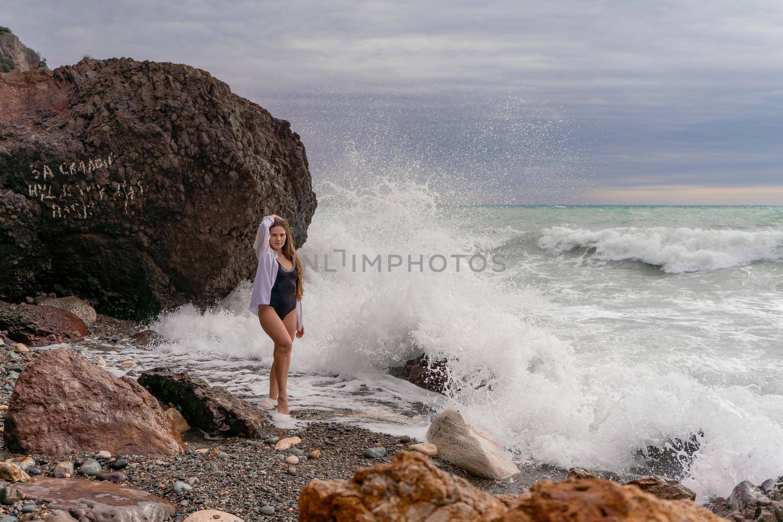 A beautiful girl in a black dress is walking on the waves, big waves with white foam. A cloudy stormy day at sea, with clouds and big waves hitting the rocks