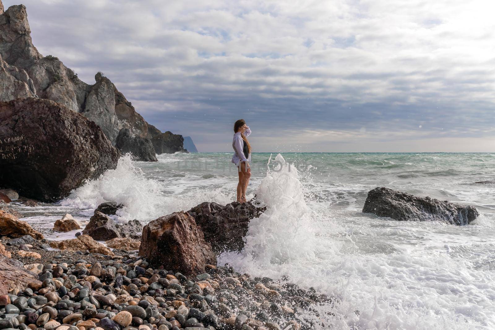 A beautiful girl in a white shirt and black swimsuit stands on a rock, big waves with white foam. A cloudy stormy day at sea, with clouds and big waves hitting the rocks