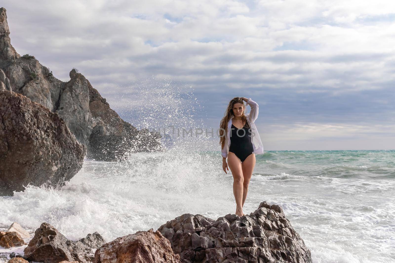 A beautiful girl in a white shirt and black swimsuit stands on the edge of a cliff. She was looking ahead. Beautiful pretty girl on the beach of the sea ocean shore with big stones. Summer outdoor lifestyle by Matiunina