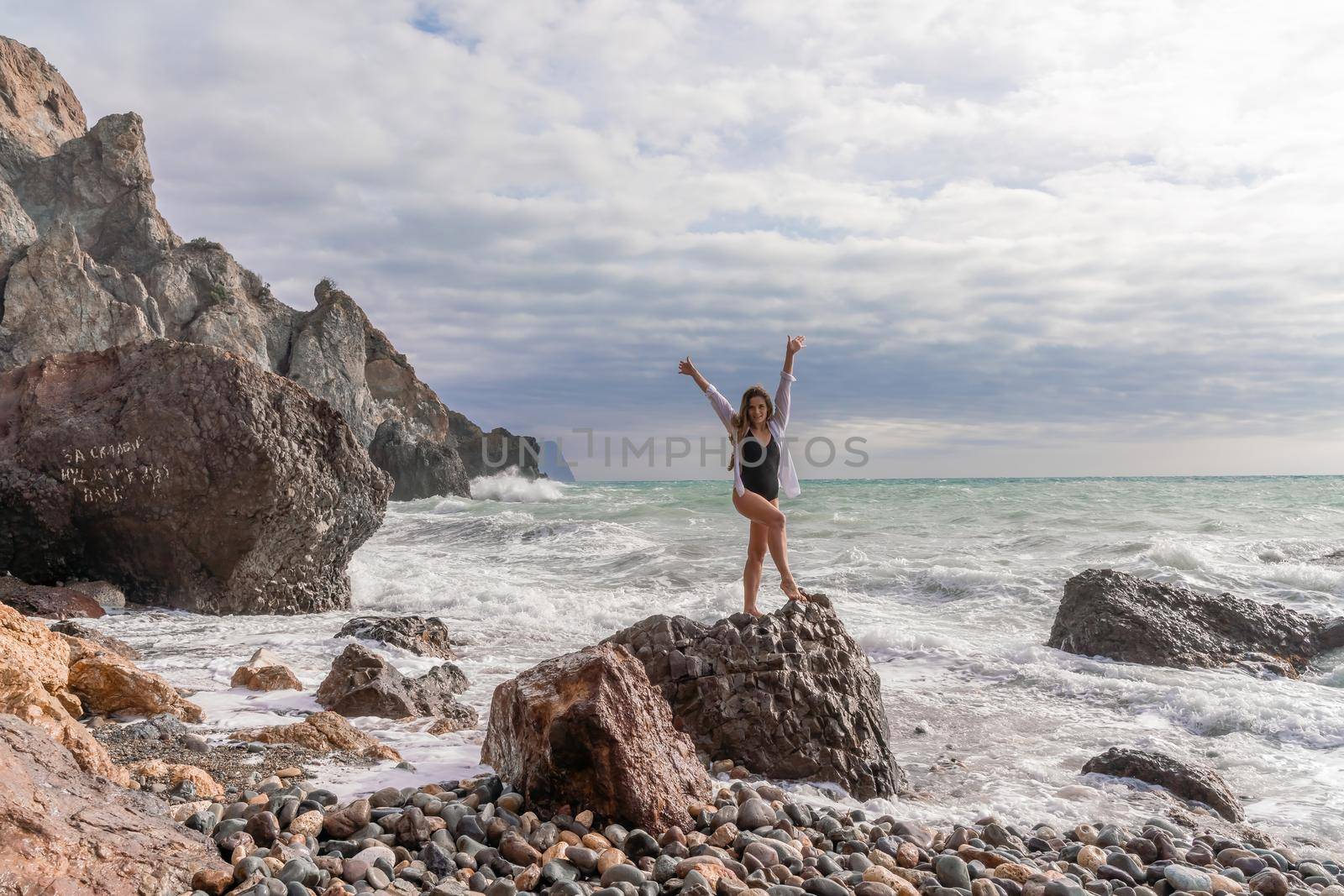 A beautiful girl in a white shirt and black swimsuit stands on a rock, big waves with white foam. A cloudy stormy day at sea, with clouds and big waves hitting the rocks. by Matiunina