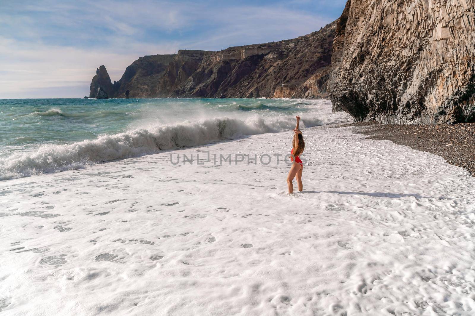 A beautiful and sexy brunette in a red swimsuit on a pebble beach, Running along the shore in the foam of the waves by Matiunina