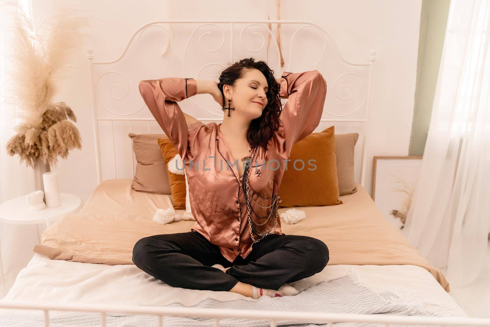 Photo of a middle aged woman in pajamas stretching her arms and smiling while sitting on the bed after sleeping or napping.