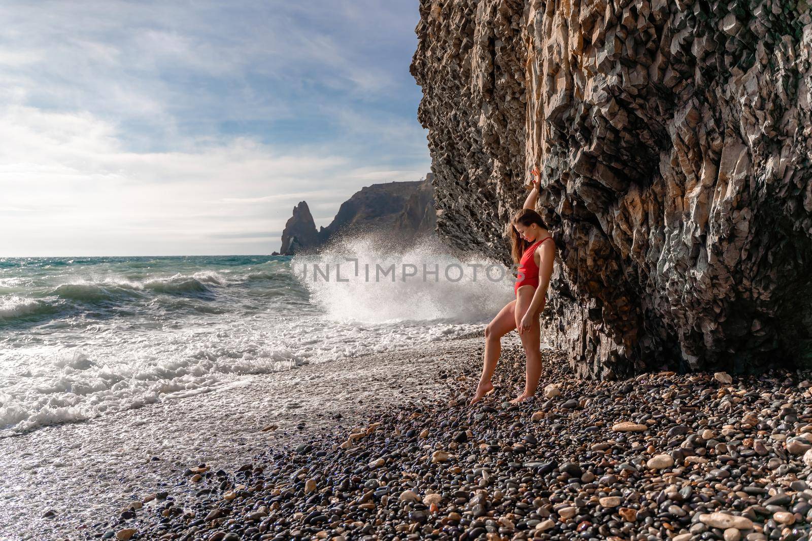 A beauty in a red swimsuit with long legs poses on a fantastic beach with huge waves against the background of mountains.