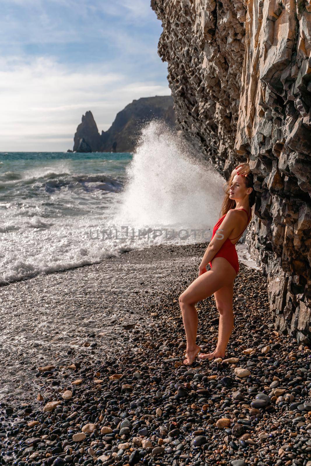 A beauty in a red swimsuit with long legs poses on a fantastic beach with huge waves against the background of mountains by Matiunina
