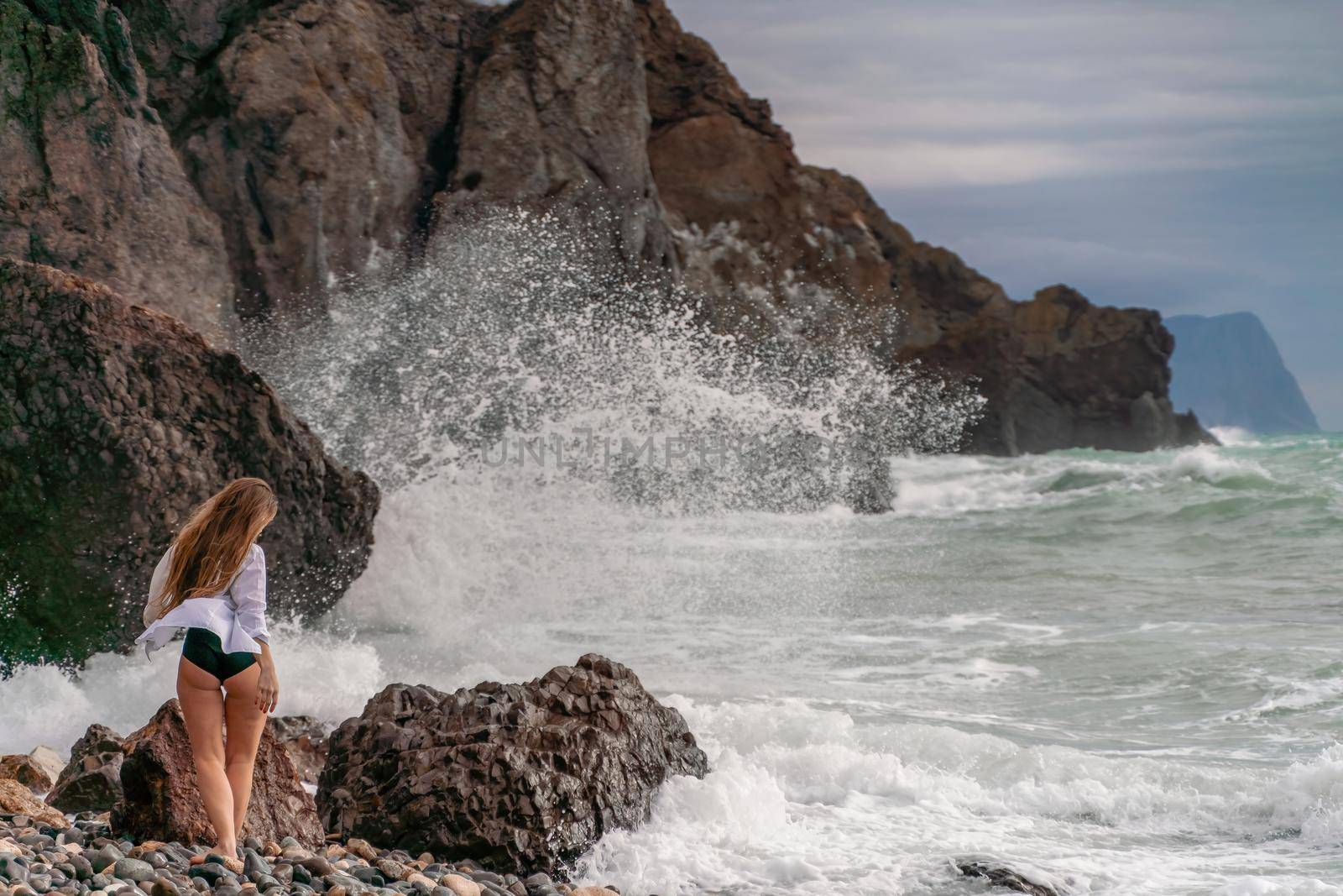 A beautiful girl in a white shirt and black swimsuit stands on the edge of a cliff, big waves with white foam. A cloudy stormy day at sea, with clouds and big waves hitting the rocks