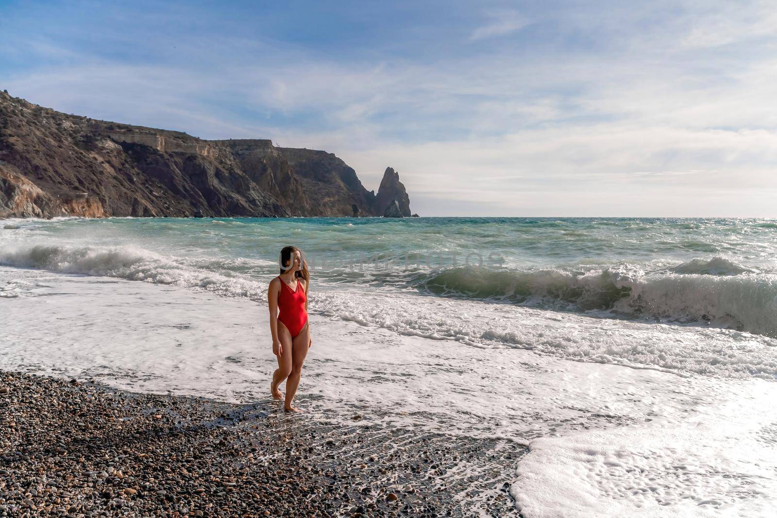 A beautiful and sexy brunette in a red swimsuit on a pebble beach, Running along the shore in the foam of the waves.
