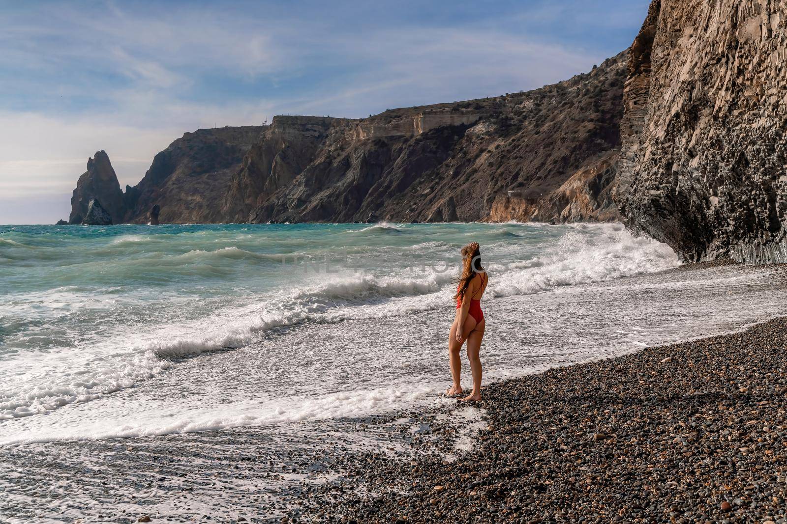 A beautiful and sexy brunette in a red swimsuit on a pebble beach, Running along the shore in the foam of the waves.