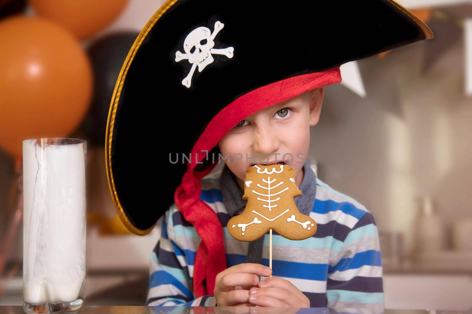 Close-up portrait of a boy dressed as a pirate for Halloween. The child eats homemade gingerbread in the form of a skeleton and drinks it with milk. House party concept for the whole family