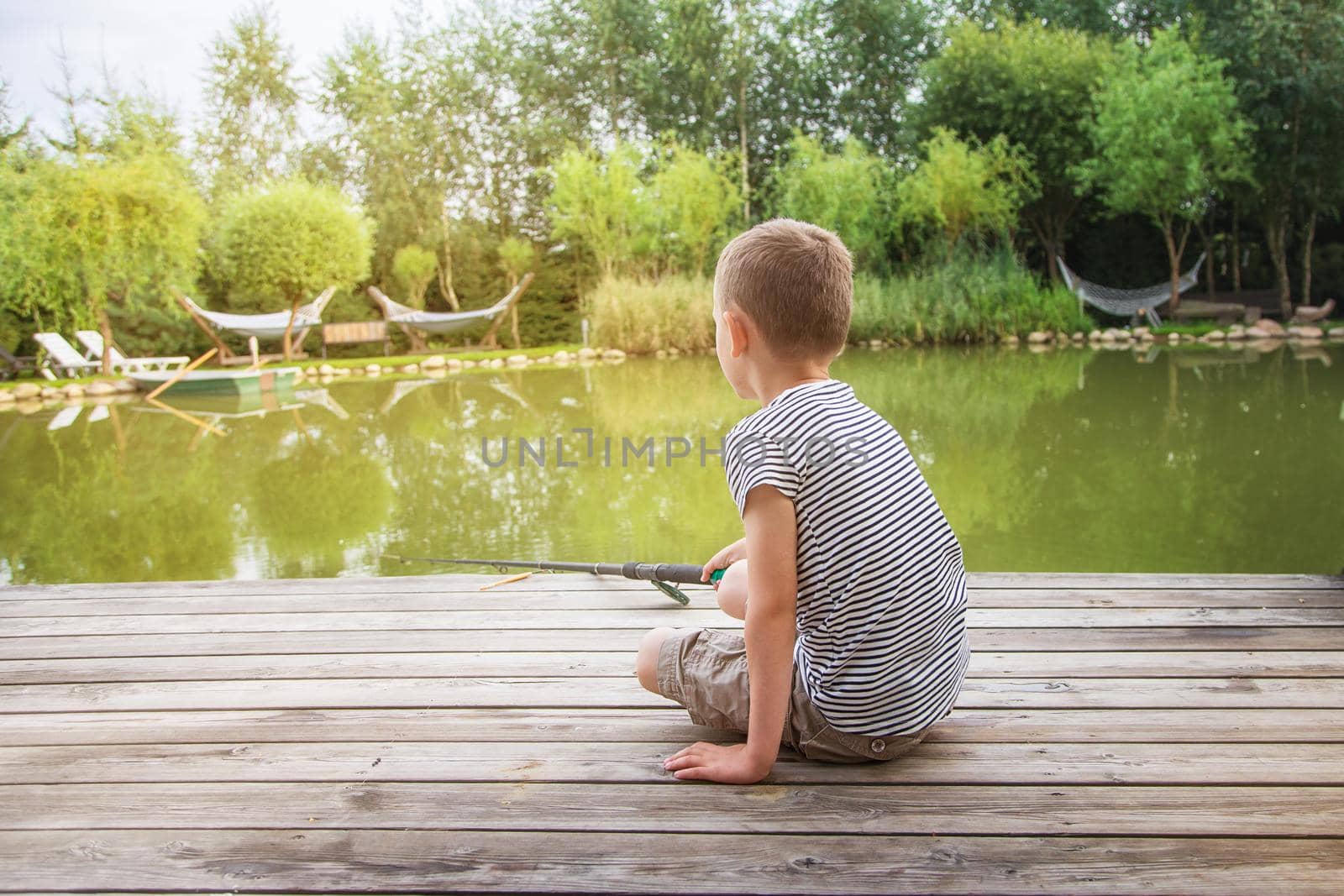 on a pier by the lake on a sunny day, a boy sits fishing. A child in a striped T-shirt and shorts sits on a wooden pier and looks into the distance, holding a fishing rod. June 1st