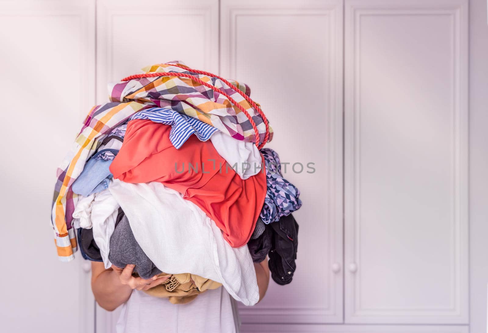 close-up, a young woman holds in her hands a large pile of colored, crumpled clothes against the background of a white wardrobe in the sun. The girl's head is not visible, copy paste for your design