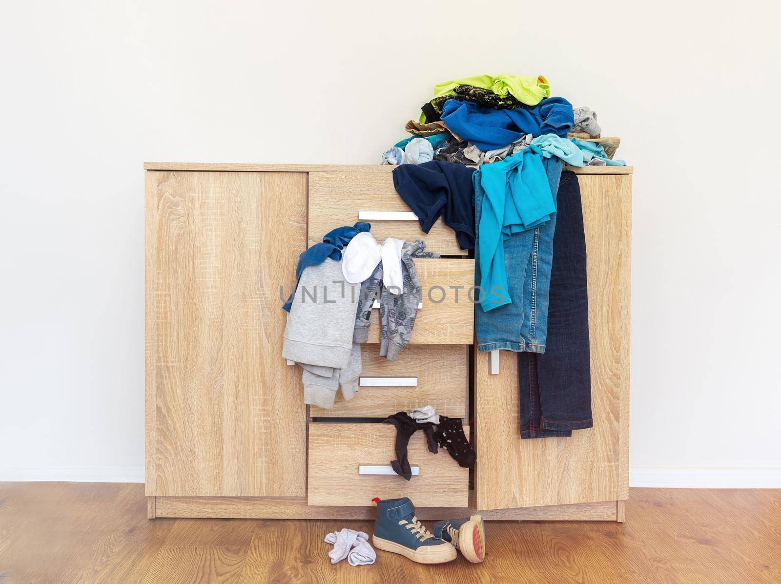 open drawers of a wooden chest of drawers with clothes falling out and piles of things around in a mess. The concept of space organization, order, home routine, cleaning. Light background, copy paste.